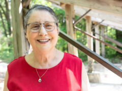Emily Kling, smiling and wearing a red t-shirt, stands near the amphitheatre in the Kreher Preserve and Nature Center with trees dotting the background.