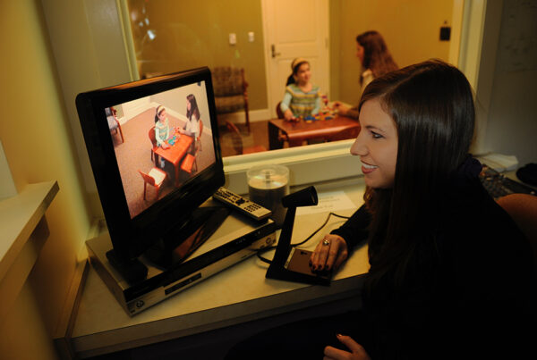A therapist watches an adult and child build blocks on a TV screen while the same child and adult can be seen through a one-way mirror in the background.
